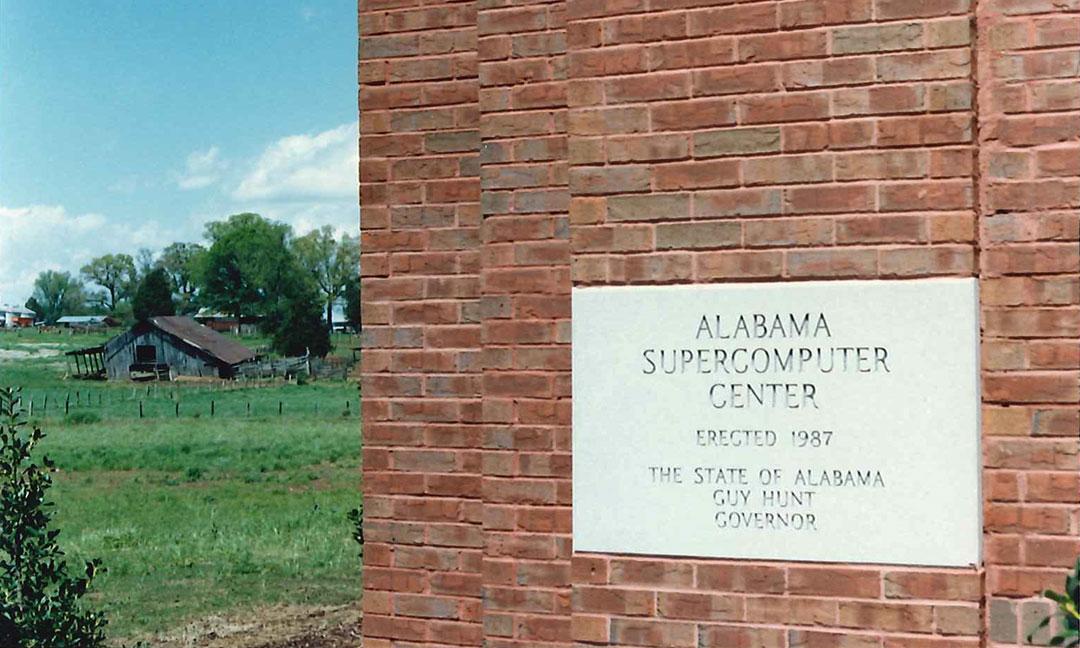 Granite cornerstone embedded in brick facade engraved with "Alabama Supercomputer Center Erected 1987 The State of Alabama Governor Guy Hunt" with old weathered barn in a green pasture in the background 