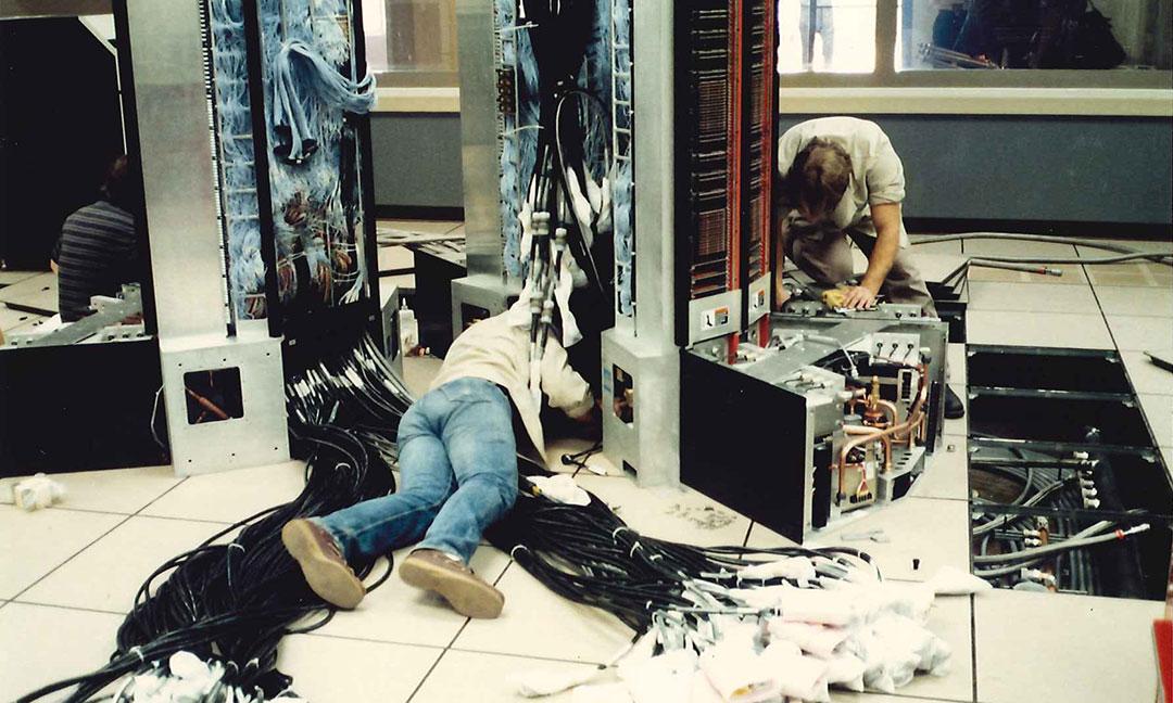 Two men working on wiring up the Cray XMP on datacenter floor with numerous bundles of wiring on the floor and removed floor tiles exposing electrical and cooling system conduits 