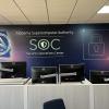 View of security operations center with rows of workstations in front of a large mural depicting the ASA logo, a SOC logo, and stylized lock icon